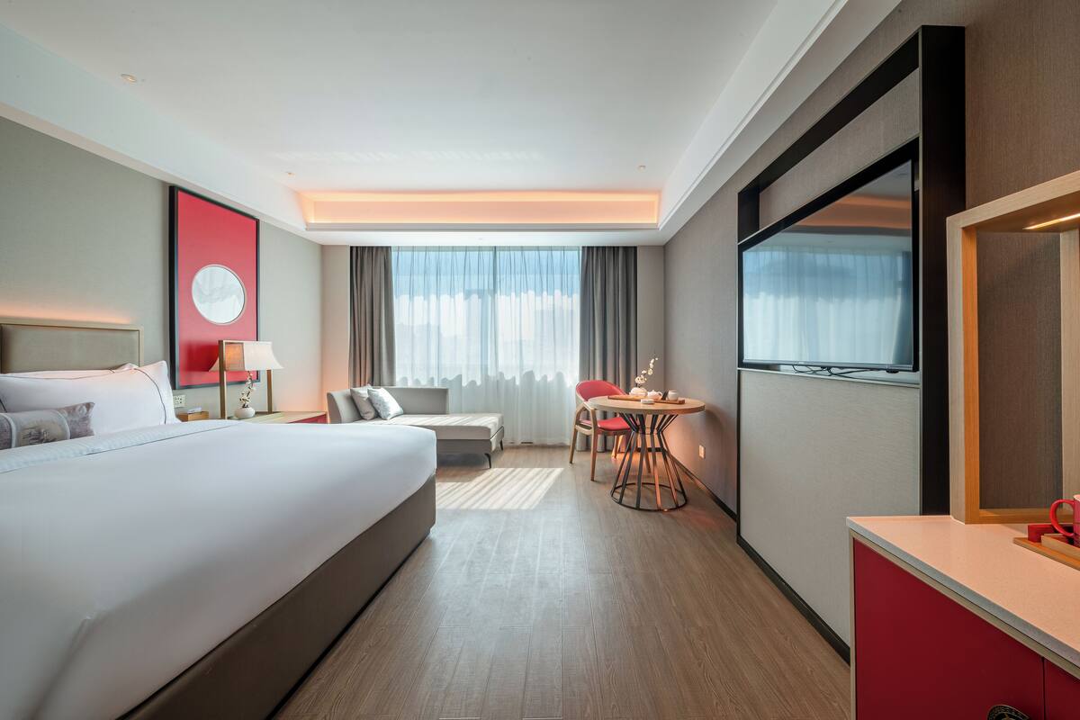 Ein Hotelzimmer mit einem großen Bett, einem Schreibtisch, einem Sessel, einem kleinen Tisch und einem Fenster mit Vorhängen.