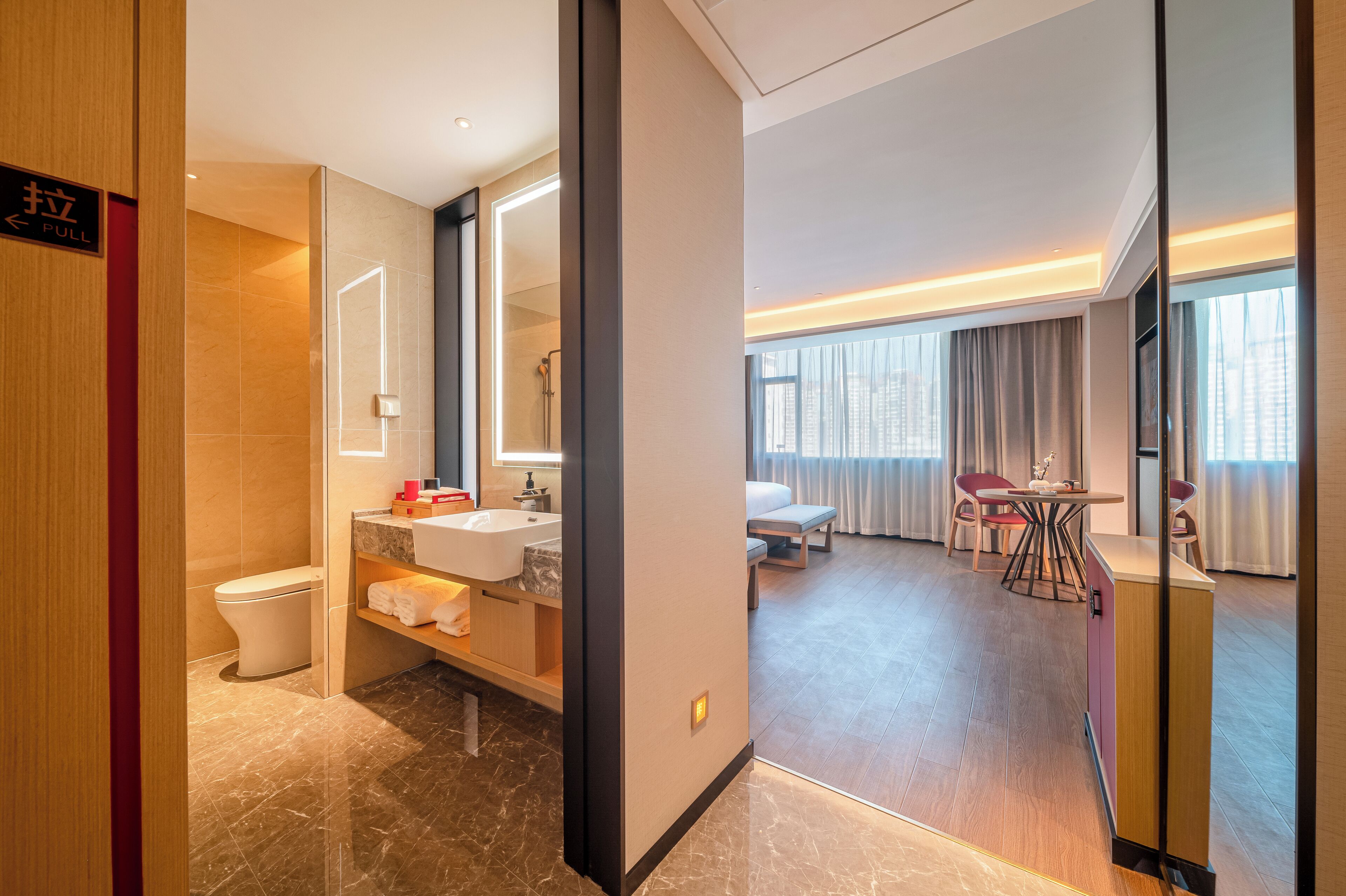 Una habitación de hotel moderna con baño, una zona de estar con mesa y sillas, y un ventanal con cortinas.