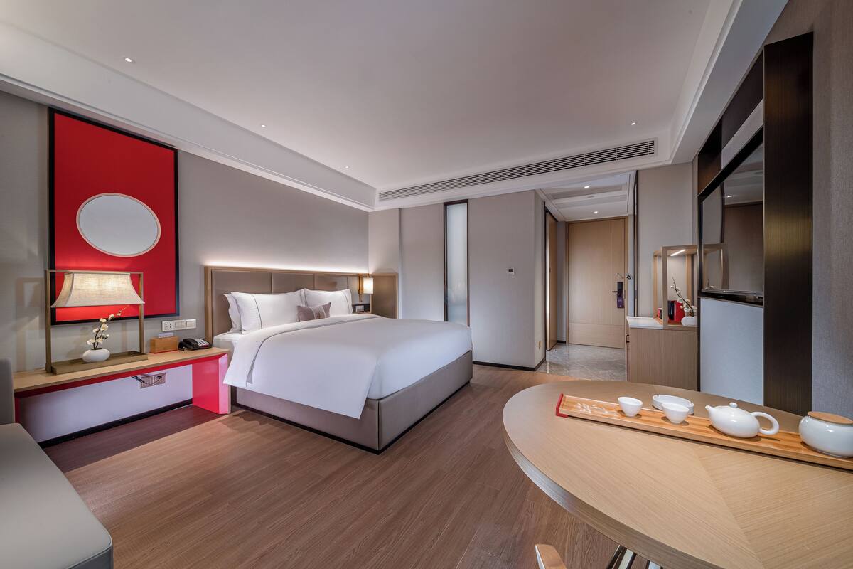 Ein modernes Hotelzimmer mit einem großen Bett, Nachttisch, Lampe und einer Frühstückstafel mit Teeset.