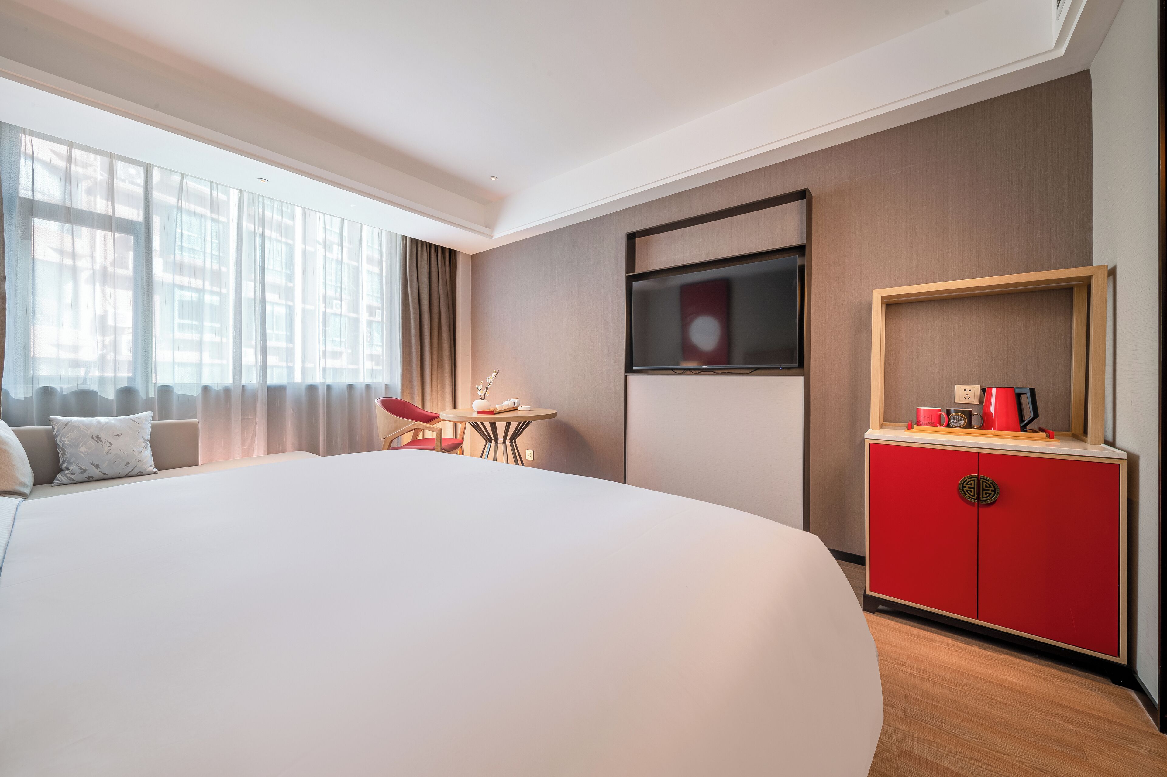 A hotel room with a large bed, a TV mounted on the wall, a small table with a chair, and a red cabinet.