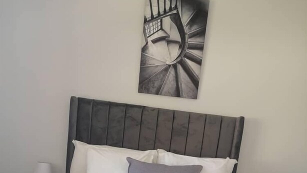 Cama bem arrumada com lençóis brancos, um travesseiro cinza e uma toalha enrolada. Há duas mesas de cabeceira com luminárias. Uma grande obra de arte abstrata está na parede acima da cama.