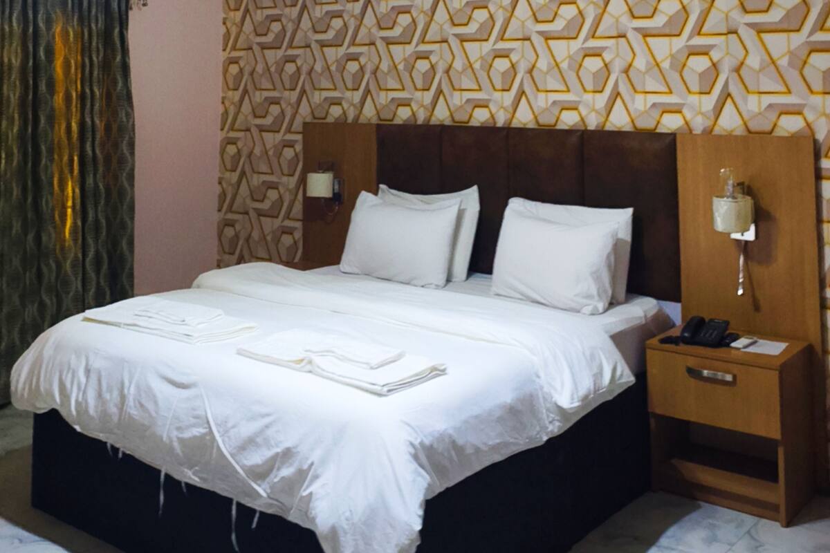 A neatly made bed with white linens and pillows, a bedside table with a lamp, and a patterned wall behind the bed.