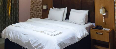 A neatly made bed with white linens and pillows, a bedside table with a lamp, and a patterned wall behind the bed.