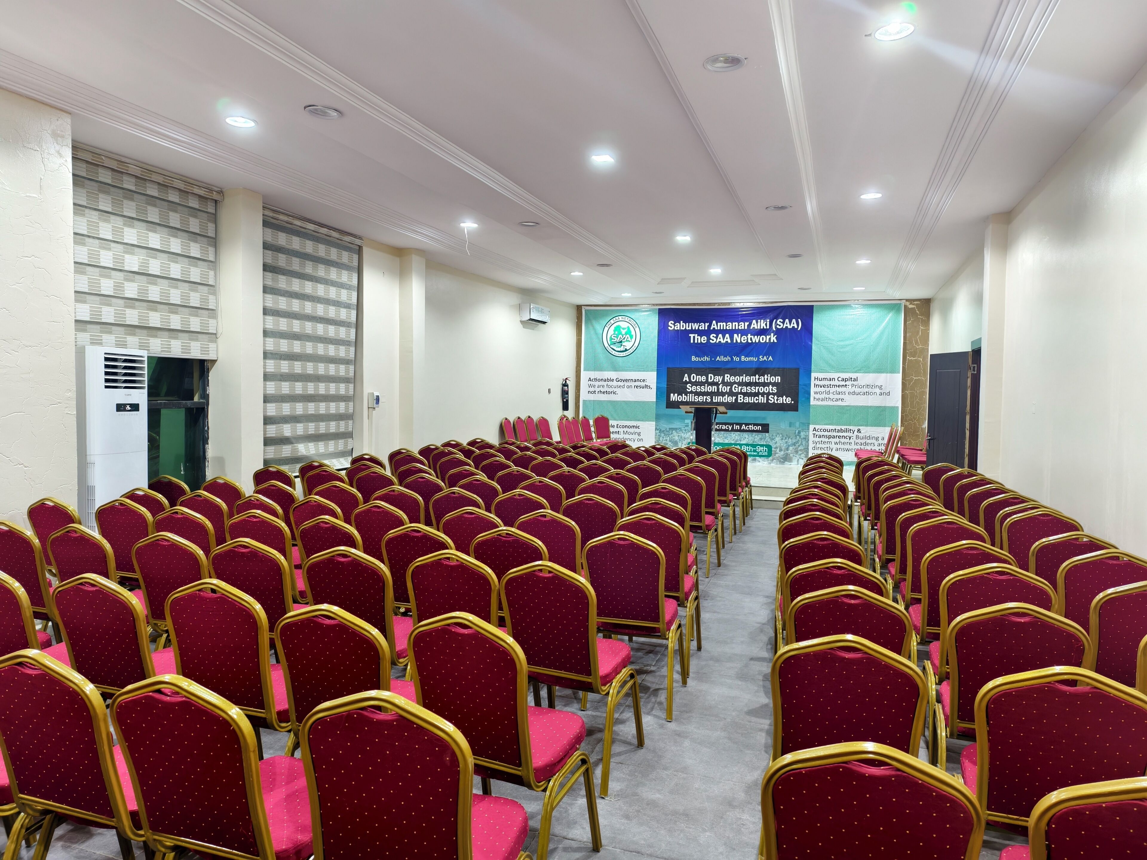 Une salle de conférence avec des rangées de chaises rouges et une scène sur laquelle est accrochée une banderole.