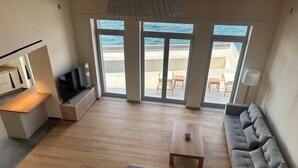 Ein modernes Wohnzimmer mit einem grauen Sofa, einem Holztisch und einem Fernseher auf einem Schrank. Große Fenster bieten Blick auf Strand und Meer.