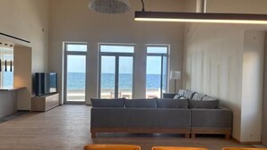 Ein geräumiges Wohnzimmer mit einem großen grauen Sofa, einem Holzesstisch und Stühlen. Große Fenster bieten Blick aufs Meer.