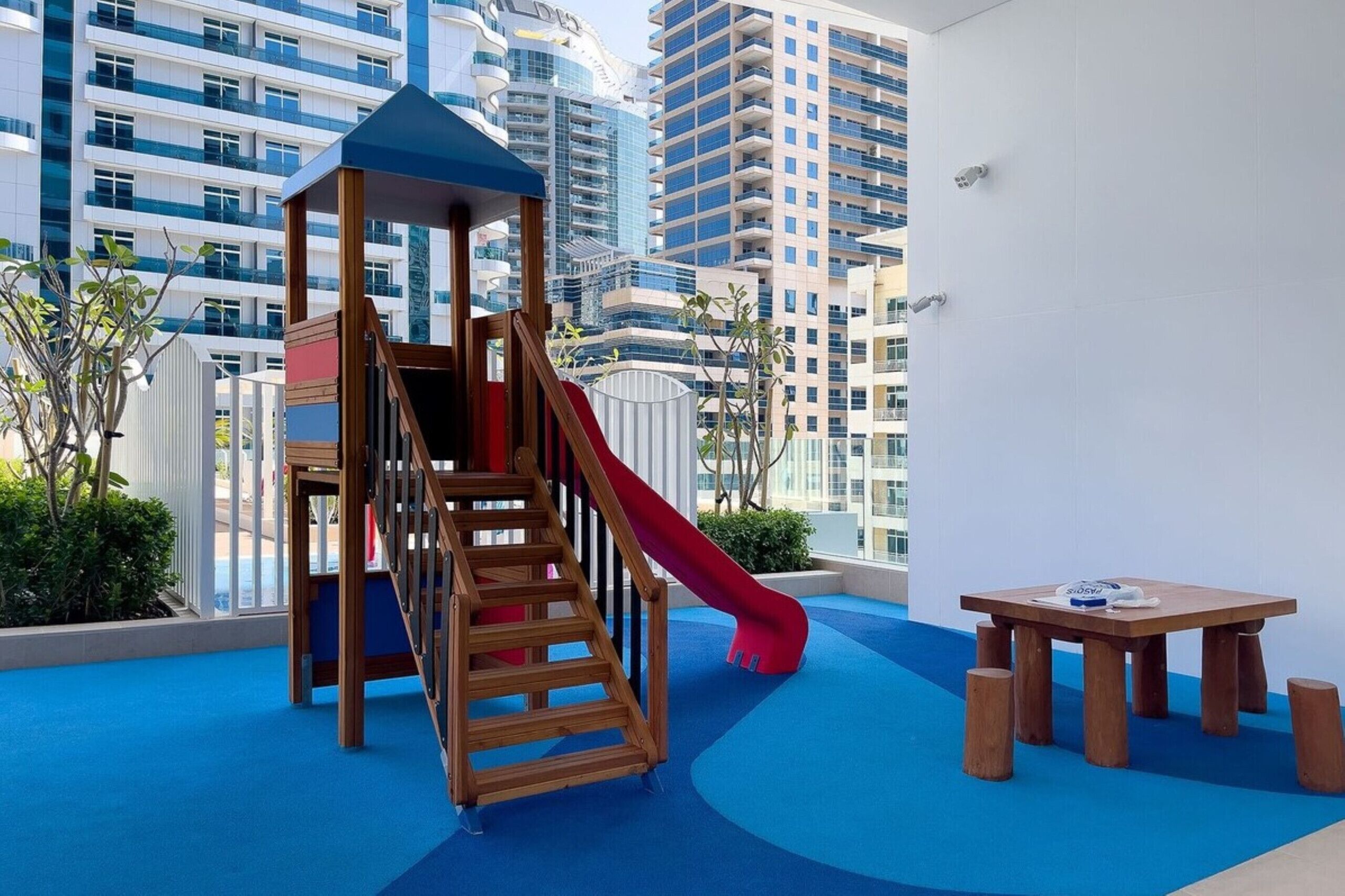 Ein Kinderspielbereich mit Rutsche und Klettergerüst aus Holz, einem kleinen Tisch und Blick auf hohe Gebäude.