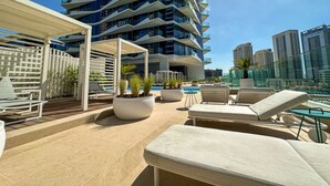Área de piscina en la azotea con sillones de descanso, jardineras y un edificio moderno al fondo.