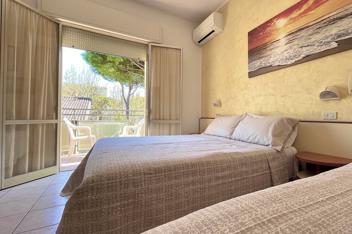 A hotel room with two beds, a balcony with chairs, and a painting of a sunset over the sea.