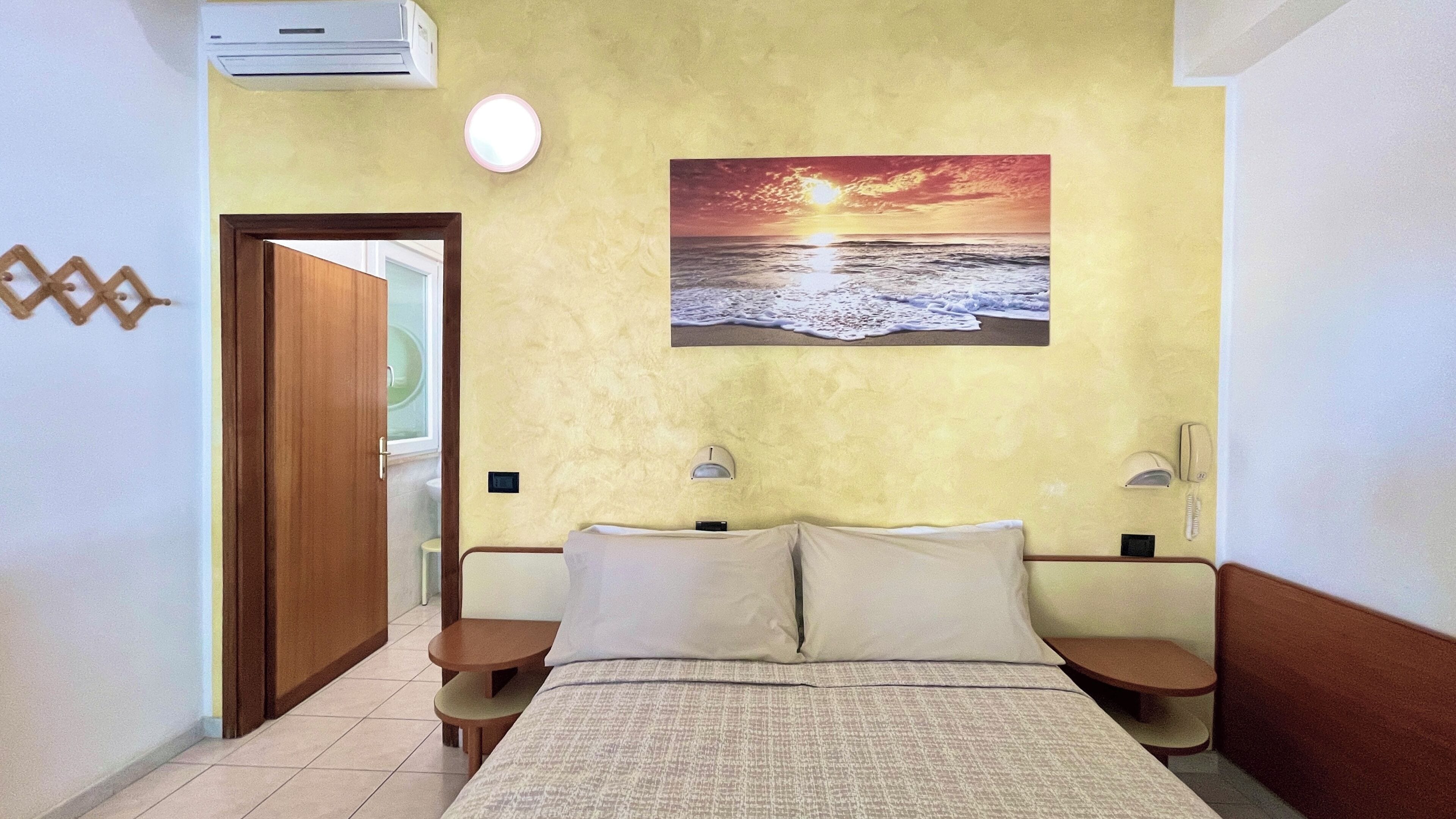 A hotel room with a bed, bedside tables, a painting of a sunset over the ocean, an air conditioning unit, and a telephone.
