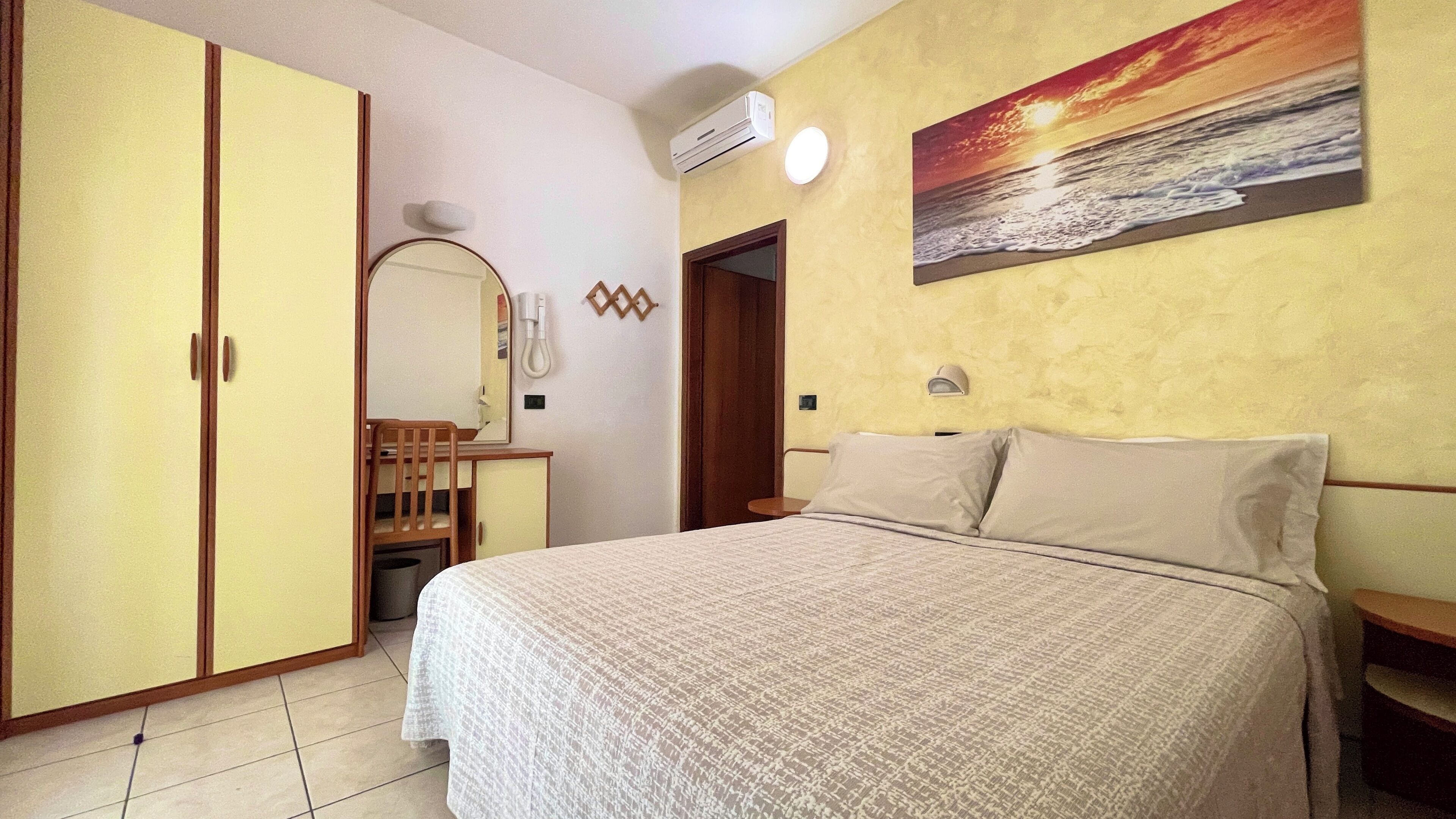 A bedroom with a bed, a desk with a chair, a wardrobe, a mirror, and a painting of a sunset over the ocean.