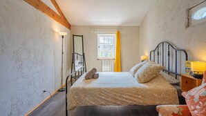Een slaapkamer met een bed, een nachtkastje, een lamp, een raam met gele gordijnen en een houten balk in het plafond.