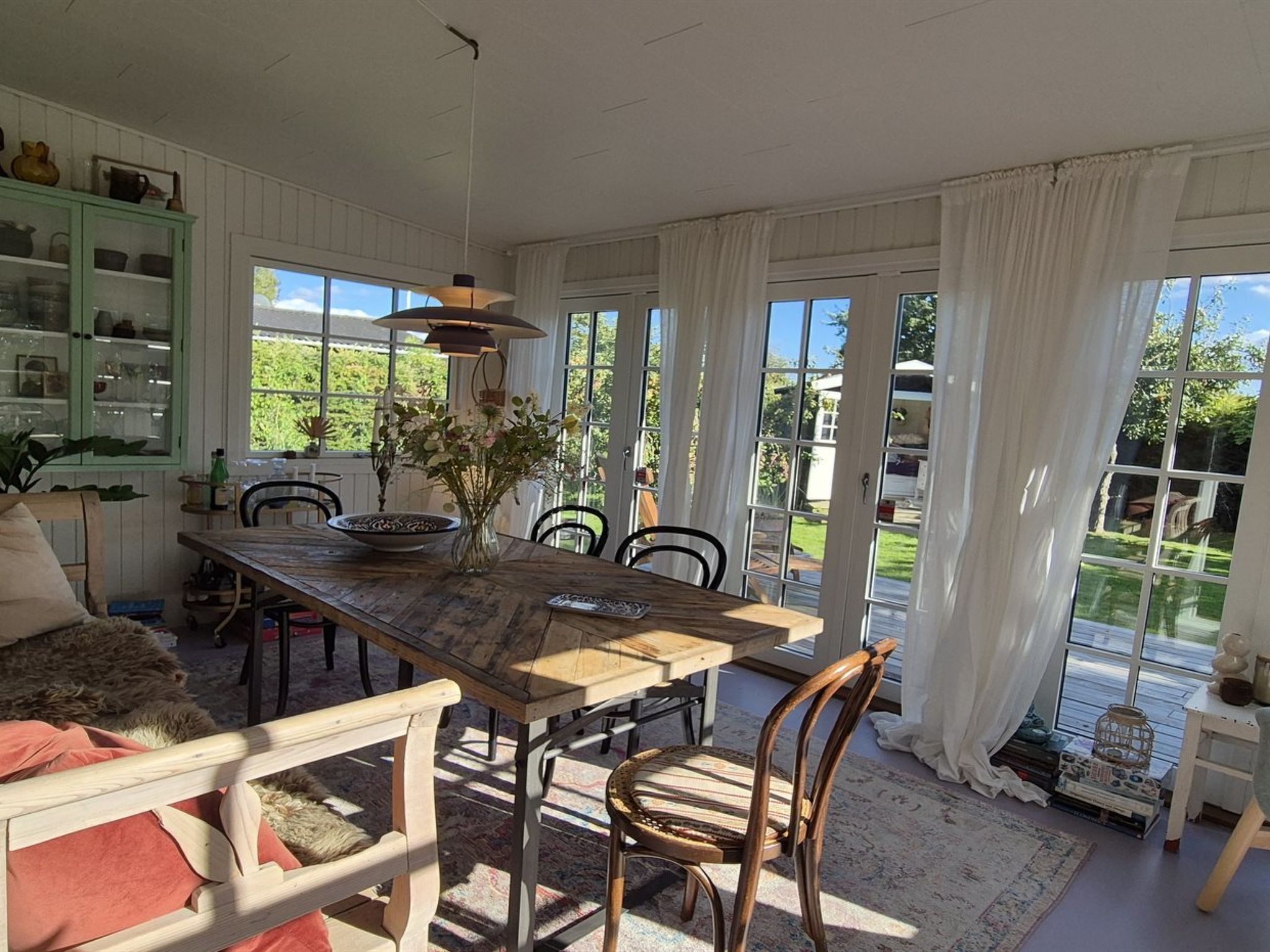 Uma sala de jantar com mesa de madeira, cadeiras e um vaso de flores. Há um armário verde com portas de vidro e vista para o exterior através de grandes janelas.