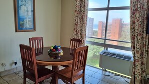 A dining area with a round table, four wooden chairs, a decorative centerpiece, and a large window with curtains. - Modern Ocean View 6 Sleeper King Suite with Wi-Fi, Underground Parking (Durban)
