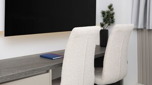 Habitación de hotel moderna con un televisor de pantalla plana grande montado en la pared, un escritorio con dos sillas blancas, una pequeña planta en maceta y una cama con ropa blanca.