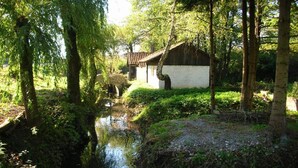 A rustic cabin with a white exterior and a brown roof, surrounded by lush greenery and a small stream.