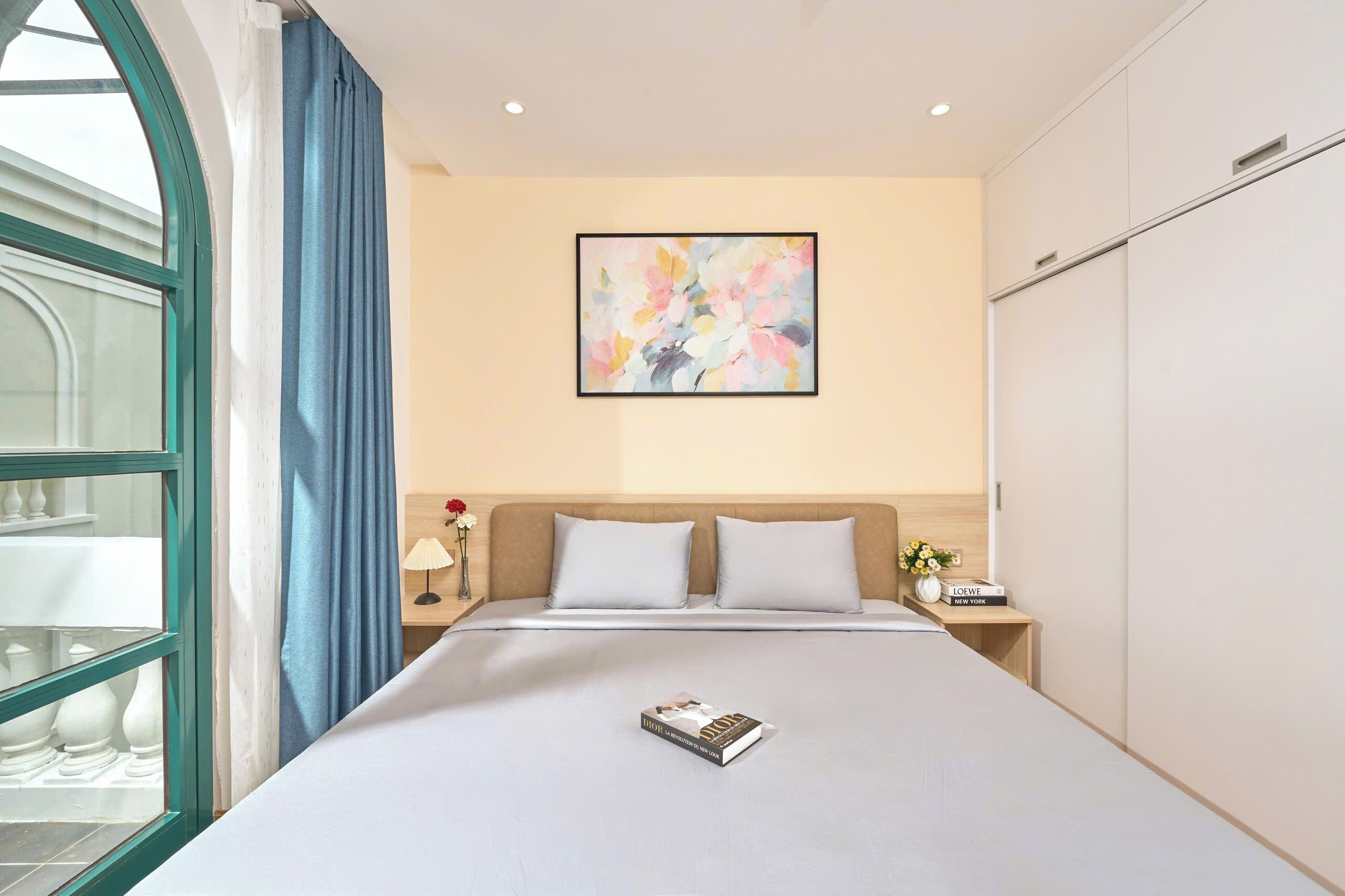 Una habitació d'hotel amb un llit gran, una finestra amb cortines blaves, un quadre abstracte penjat a la paret i una taula de nit amb una làmpada i flors.