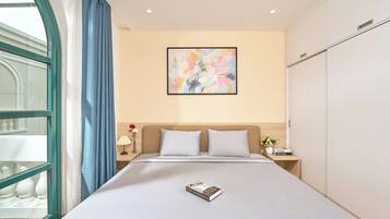 Una habitació d'hotel amb un llit gran, una finestra amb cortines blaves, un quadre abstracte penjat a la paret i una taula de nit amb una làmpada i flors.