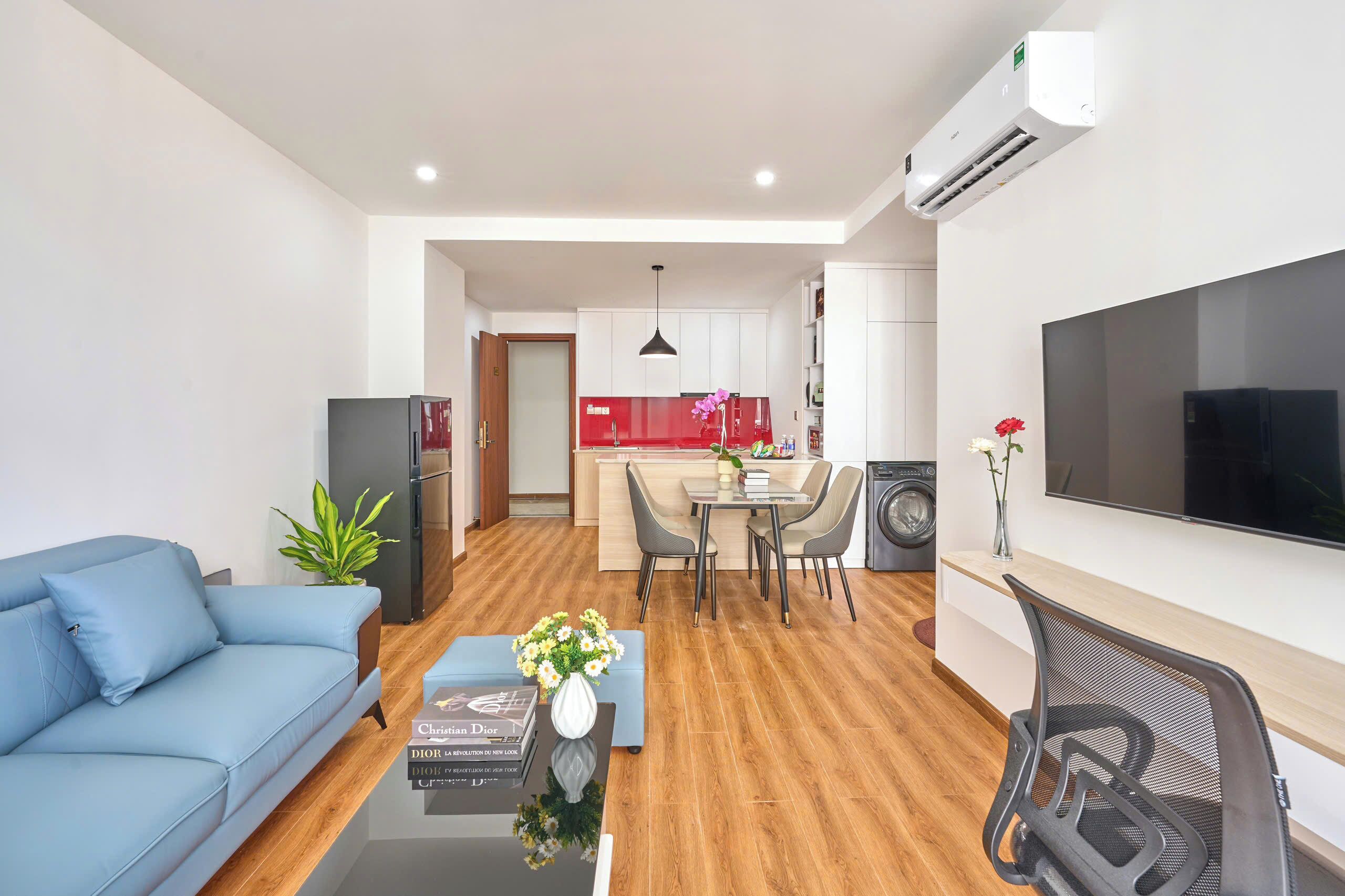A modern living room with a blue sofa, a coffee table with a book and flowers, a dining area with a table and chairs, a kitchen with a red backsplash, and a TV mounted on the wall.