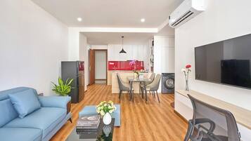 A modern living room with a blue sofa, a coffee table with a book and flowers, a dining area with a table and chairs, a kitchen with a red backsplash, and a TV mounted on the wall.