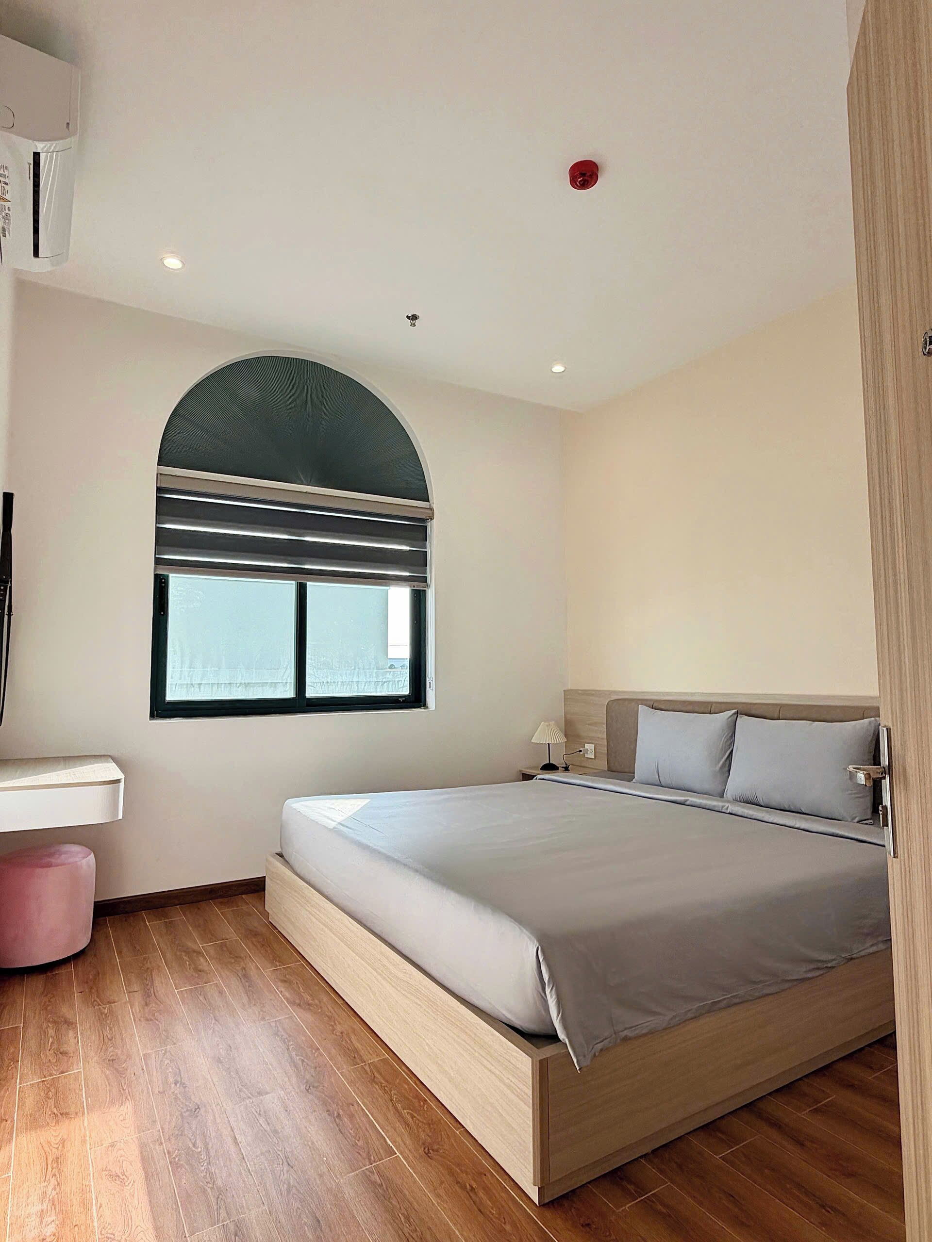 A modern bedroom with a large bed, a window with blinds, a bedside table, and a wooden floor.