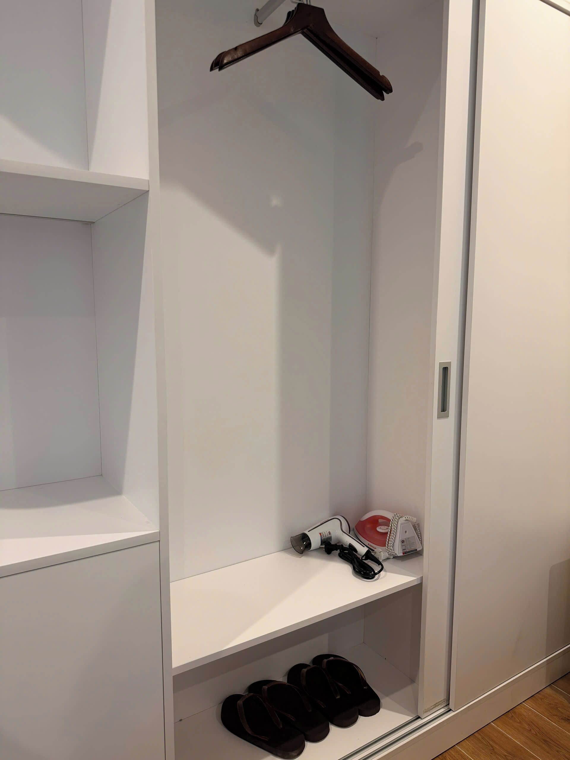 A white wardrobe with a hanging rod, a shelf with a hairdryer, and a shelf with black slippers.
