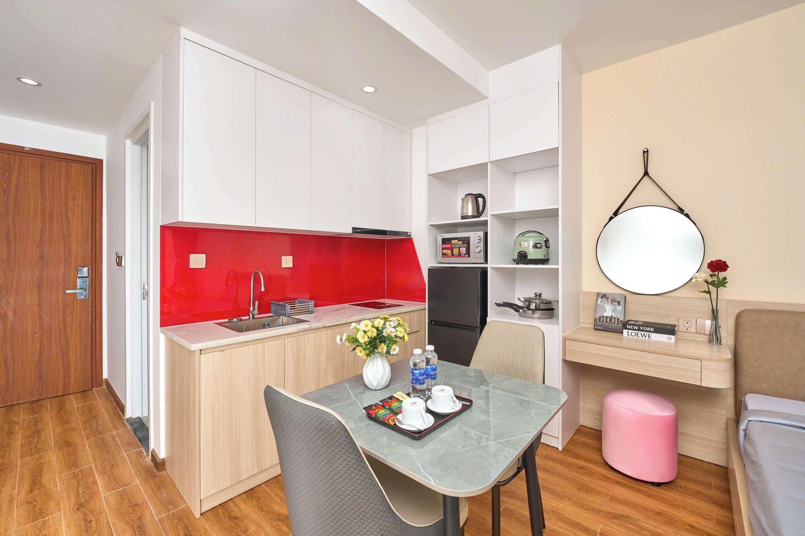 A compact kitchen with a red backsplash, a dining area with a table and chairs, a small pink stool, and a wall-mounted mirror.