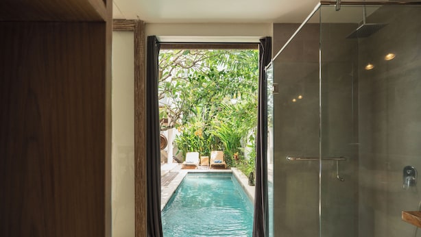 Zona de ducha con paredes de vidrio al lado de una piscina, con vistas a un área de estar exterior y vegetación.