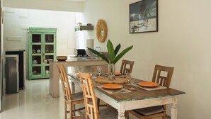 Un comedor con una mesa de madera dispuesta para cuatro personas, rodeada de sillas. Se observa una cocina con un mueble verde y un refrigerador al fondo.