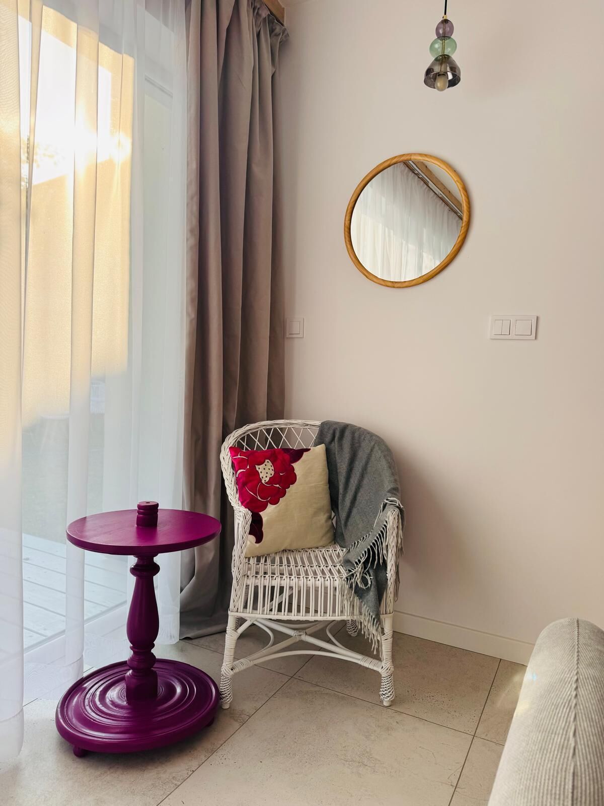 A room with a white wicker chair, a round mirror, a small round table, and a window with curtains.