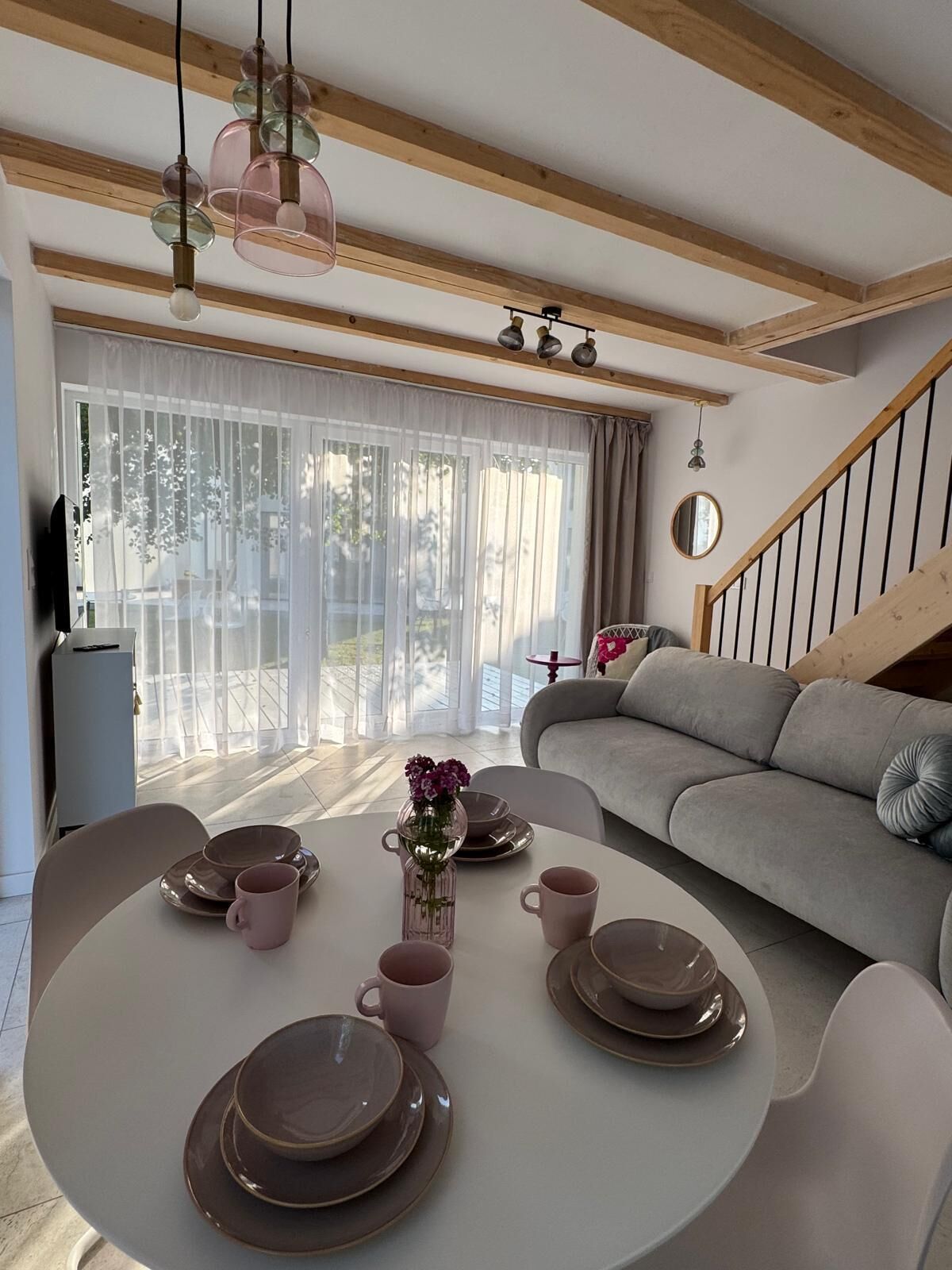 A modern living room with a round dining table set for four, a grey sofa, and a staircase.