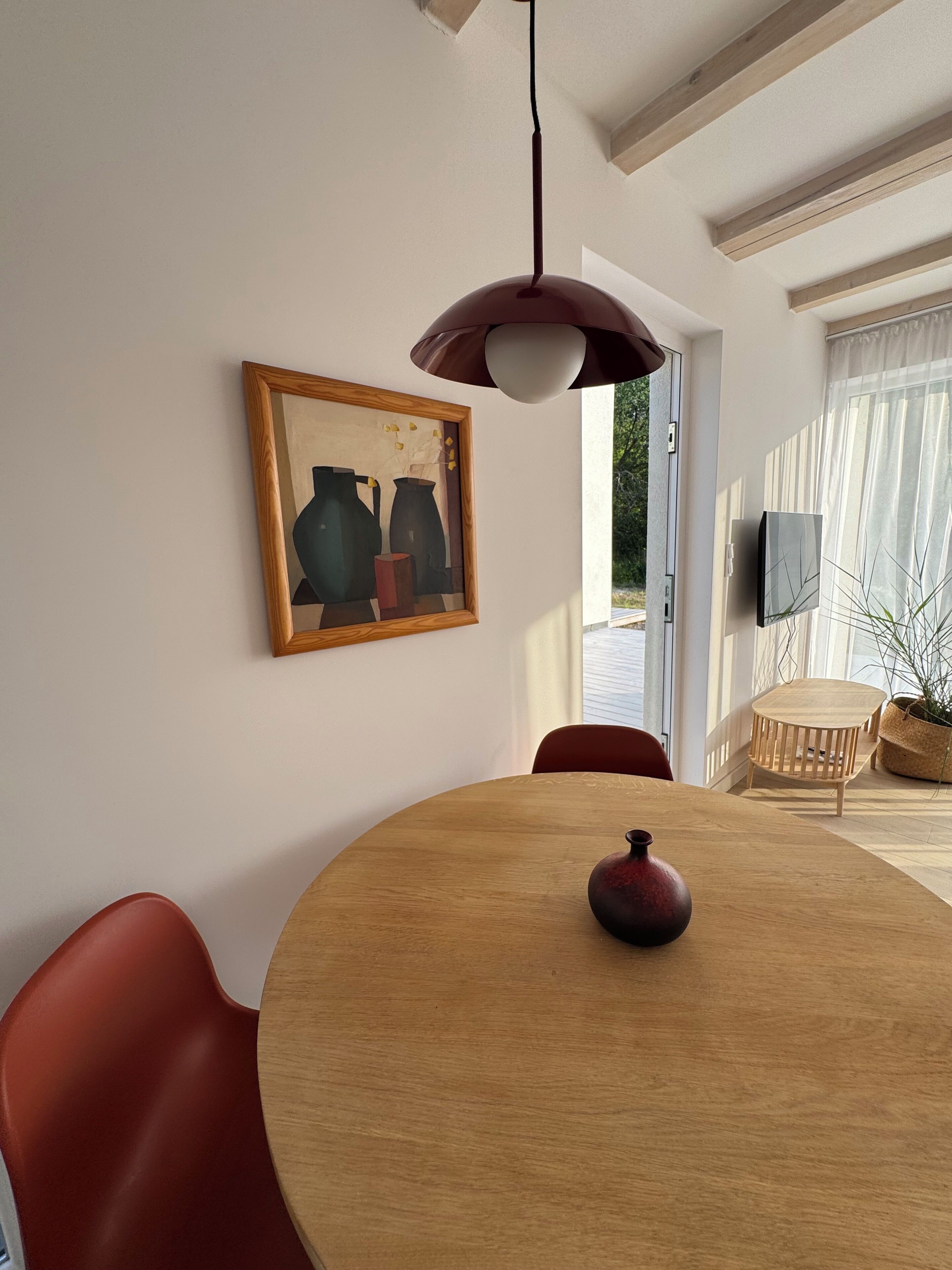 A dining area with a round wooden table, red chairs, a vase, a pendant light, and a framed artwork on the wall.