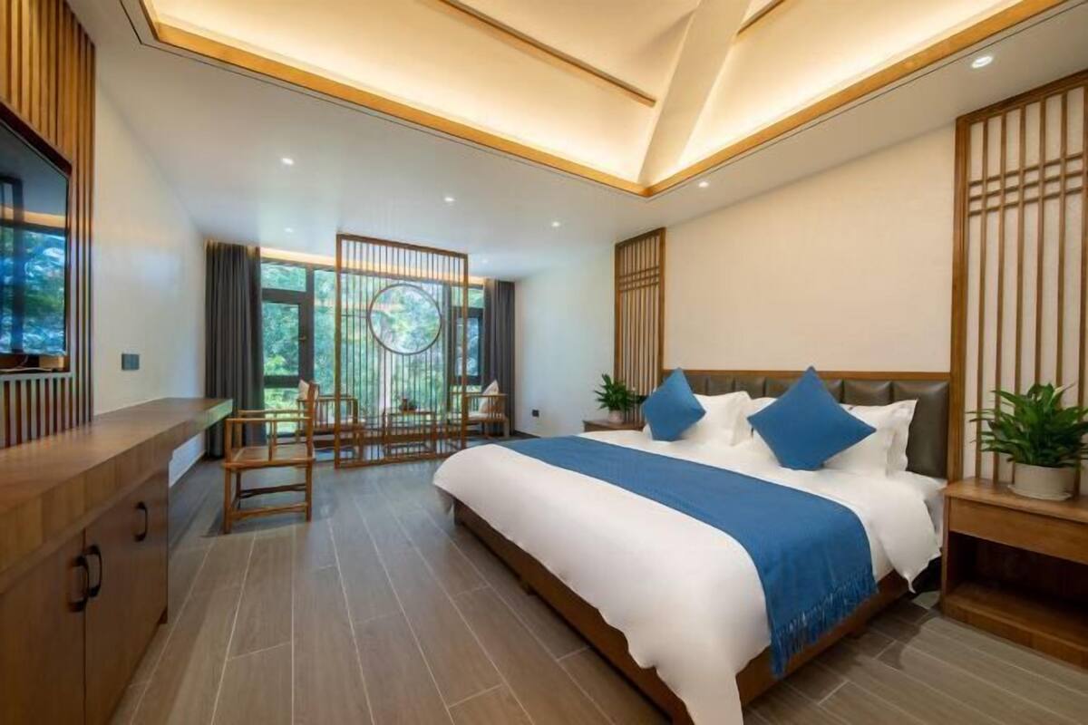 A modern bedroom with a large bed, a wooden desk, and a view of greenery outside.