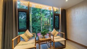 A room with three wooden chairs, a small table, and a view of a forest through large windows. - Babaling Leisure Hostel (Hengyang)