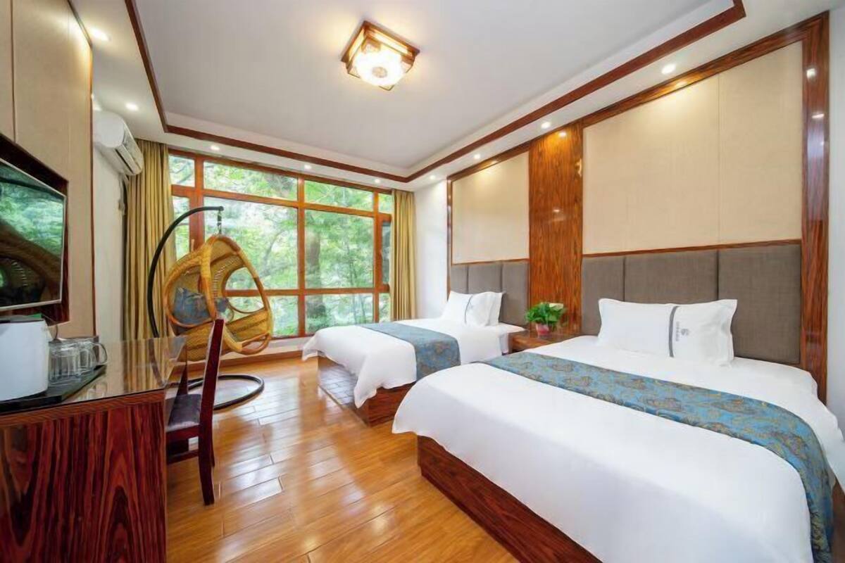 A hotel room with two beds, a wooden floor, a TV, and a large window with a view of trees.