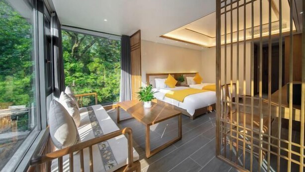A modern bedroom with a large window, a bed with white bedding and yellow accents, a wooden coffee table, and a view of greenery outside. - Babaling Leisure Hostel (Hengyang)