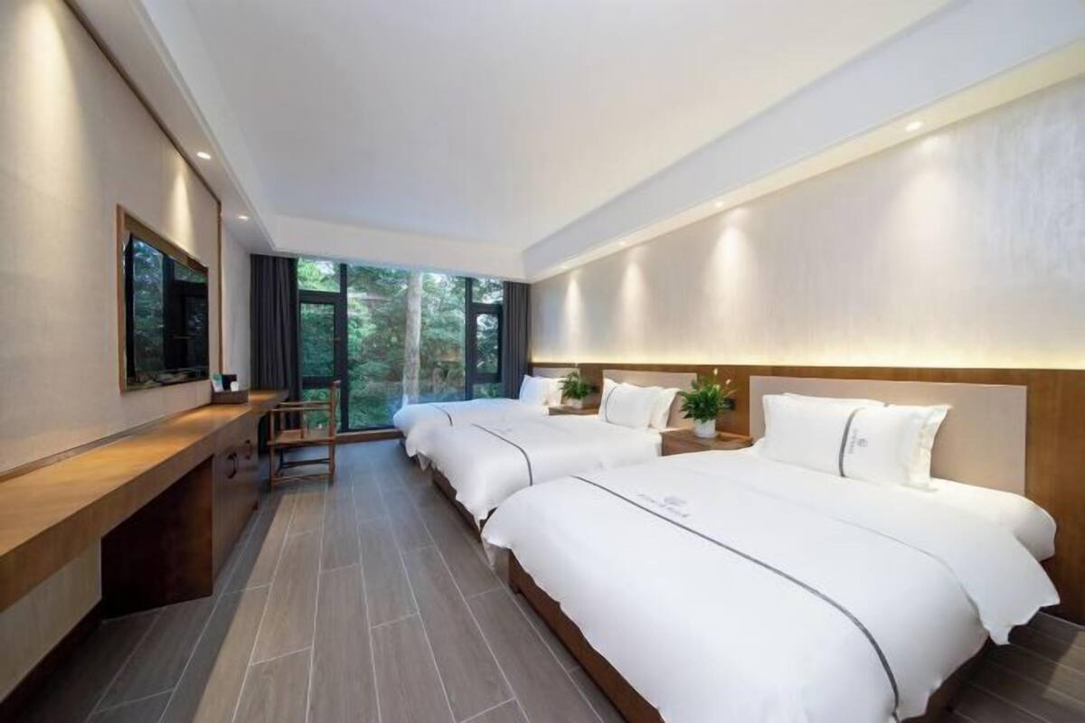 A modern hotel room with two beds, a wooden bench, a large window, and a flat-screen TV.