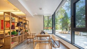 A modern interior with a wooden bookshelf, a dining table with chairs, and large windows offering a view of the outdoors. - Babaling Leisure Hostel (Hengyang)