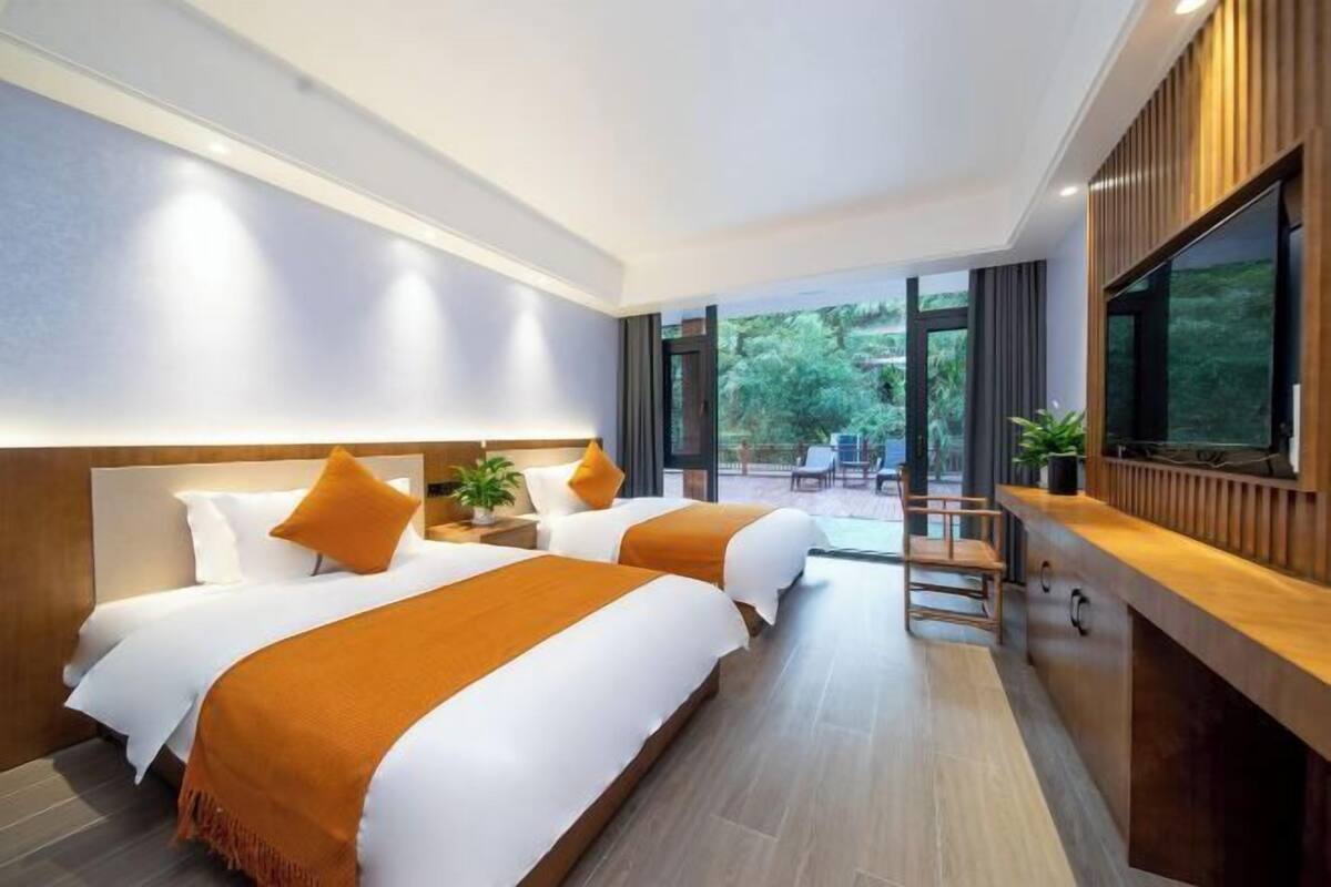 A hotel room with two beds, a wooden desk, a TV, and a balcony with a view of greenery.