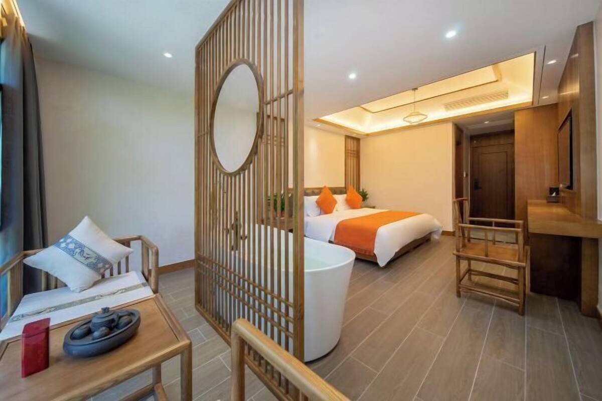 A hotel room with a bed, a desk, a chair, a small table with a bowl and a red object, and a partition with a mirror.