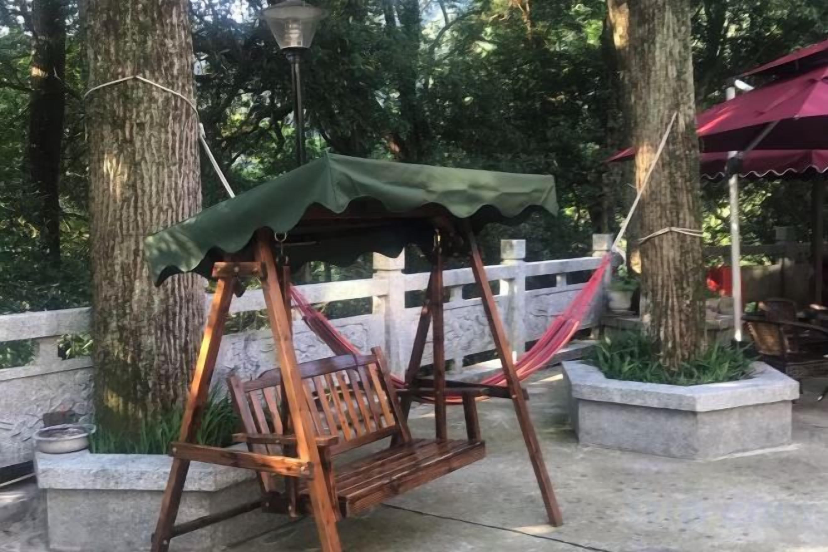 A wooden swing with a green canopy, situated in a park-like setting with trees and a white fence in the background.