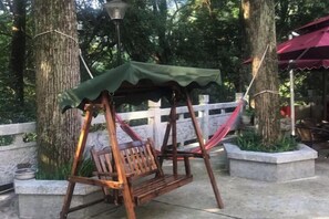 Une balançoire en bois surmontée d’une sorte de toit vert, installée dans un espace aménagé comme un parc, avec des arbres et une clôture blanche en arrière-plan.