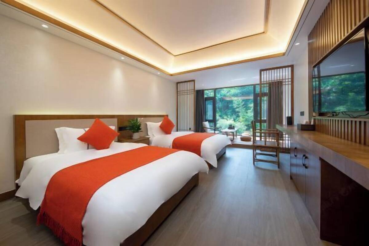 A modern hotel room with two beds, a wooden desk, and large windows offering a view of greenery.