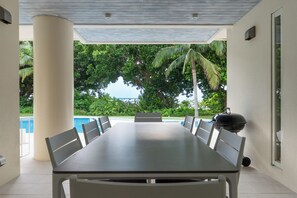 Een terras met een eettafel en stoelen, een barbecue en uitzicht op een zwembad en bomen.