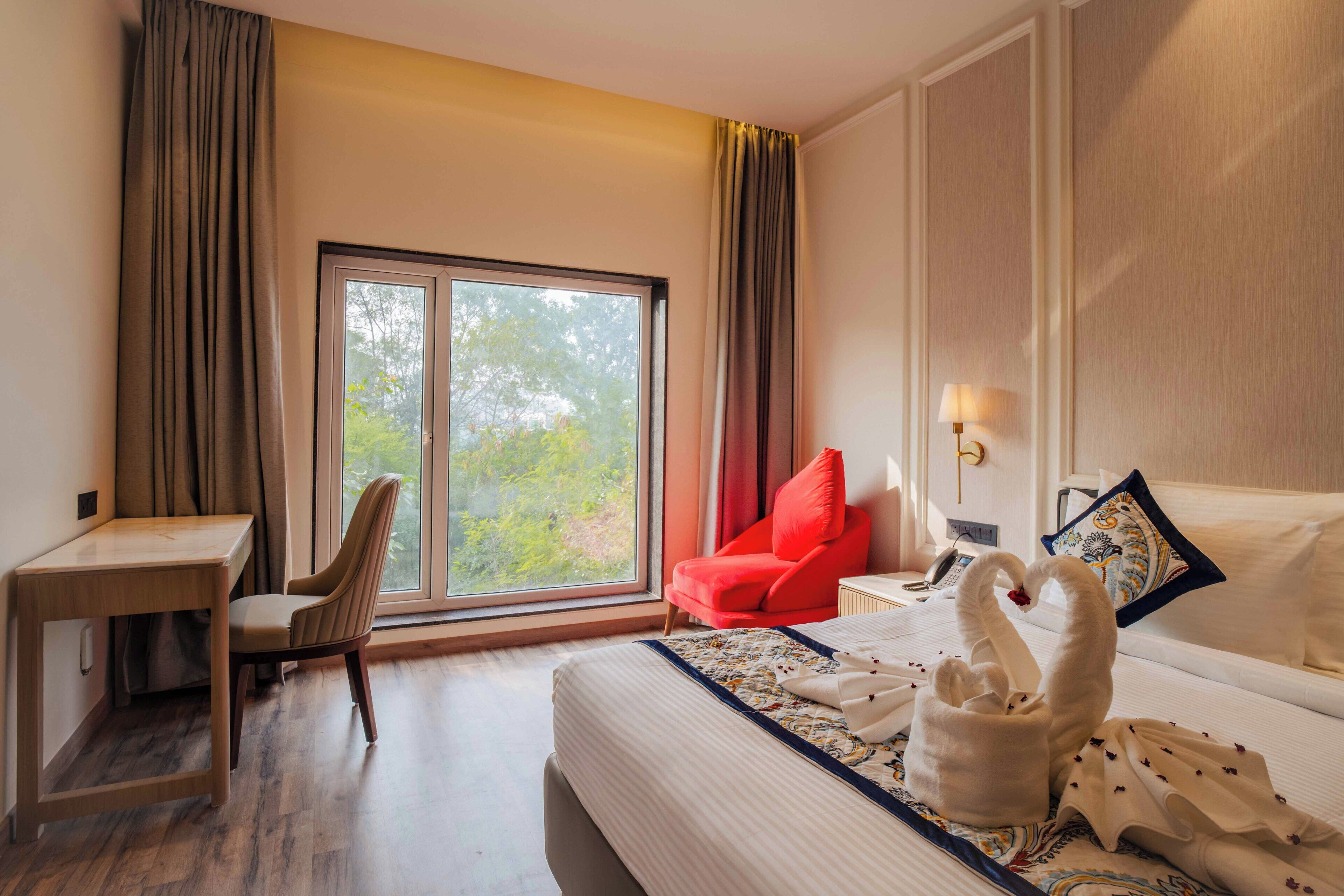 A hotel room with a bed, a desk, a chair, a window with curtains, and a lamp.