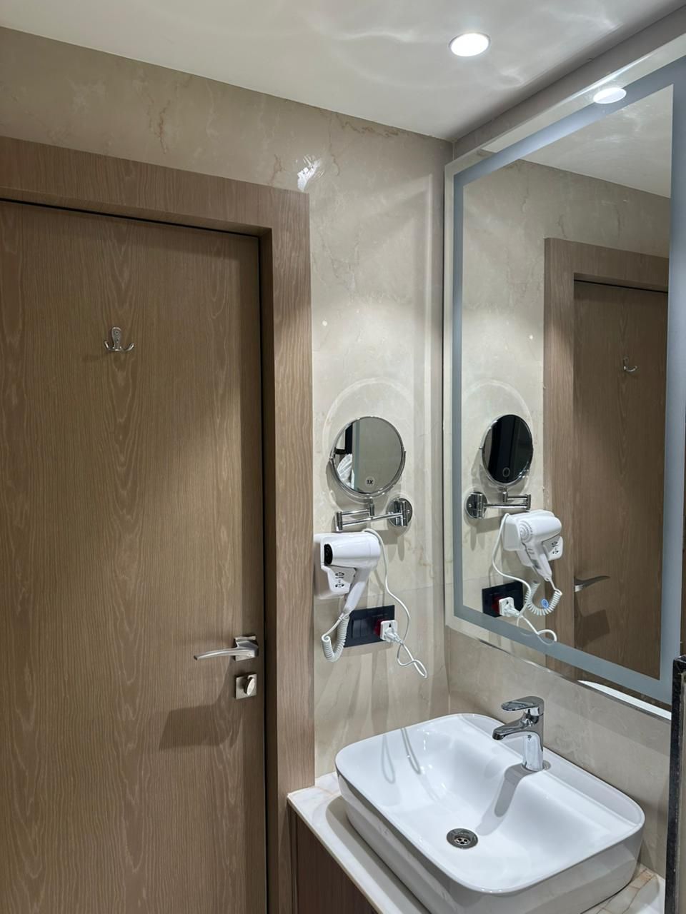 Een moderne badkamer met een houten deur, een grote spiegel, een witte wastafel en twee haardrogerapparaten bevestigd aan de muur.