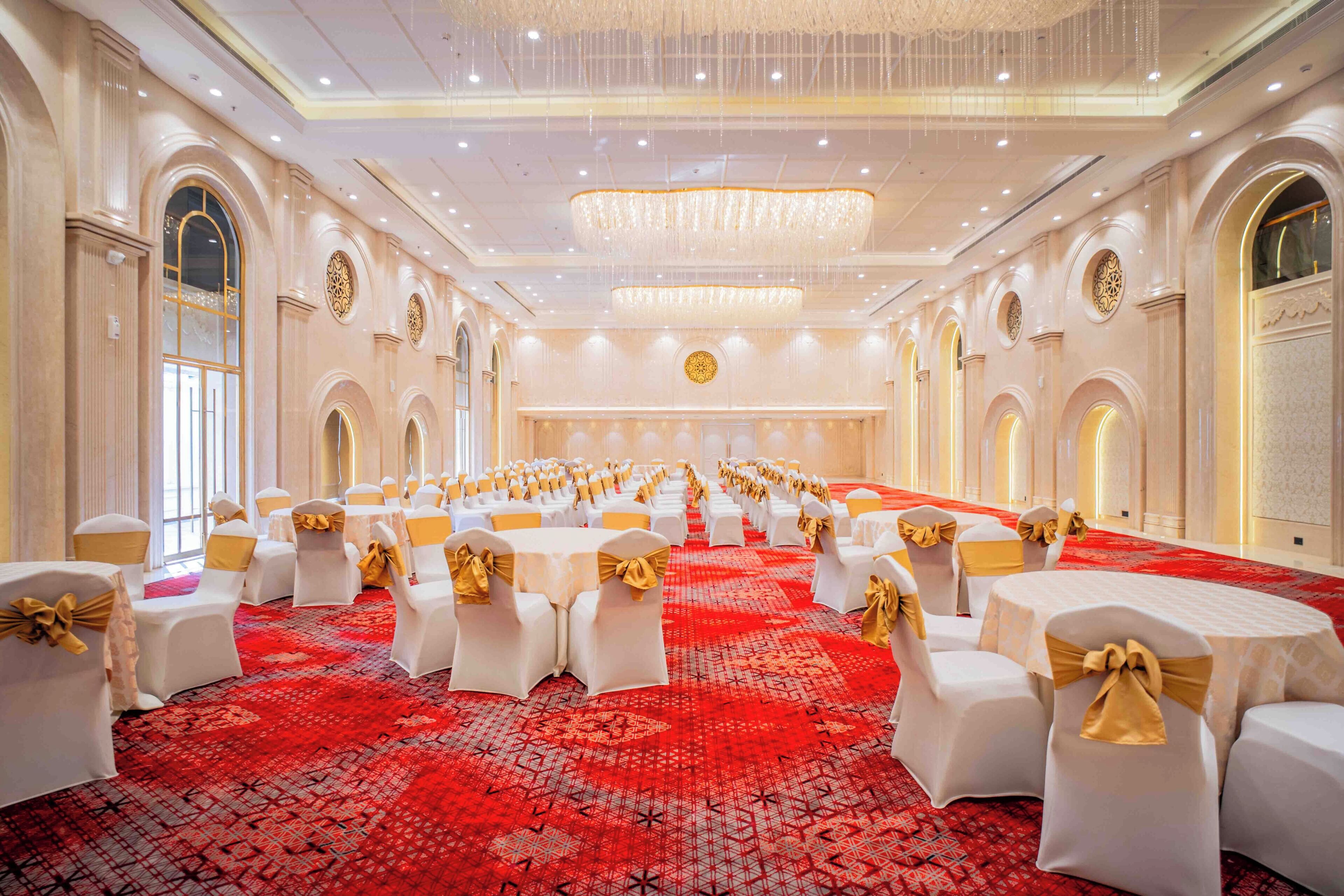 Un salón de banquetes con mesas redondas, sillas blancas con cintas doradas y una alfombra roja con motivos.