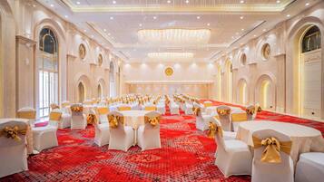 Un salón de banquetes con mesas redondas, sillas blancas con cintas doradas y una alfombra roja con motivos.