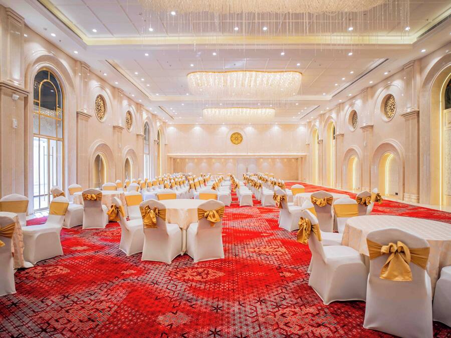 A banquet hall with round tables, white chairs with gold sashes, and a red patterned carpet.