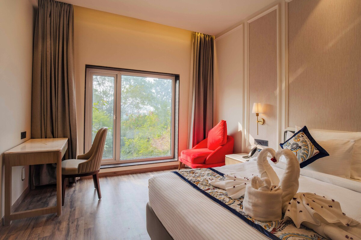 A hotel room with a bed, a desk, a chair, a window with curtains, and a lamp.